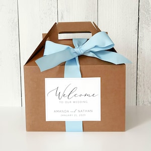 May include: A brown cardboard gift box with a blue ribbon and a white label that says "Welcome to our wedding Amanda and Nathan January 21, 2023".