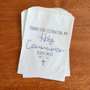 May include: White paper favor bag with the text "Thanks for celebrating my Holy Communion Olivia Smith April 2, 2022" and a purple cross.