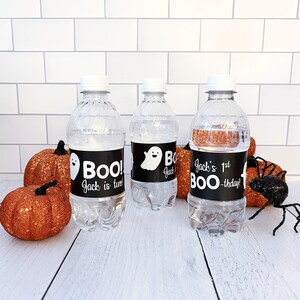 May include: Three clear plastic water bottles with white caps and black labels that say "Boo! Jack is two", "Boo!" and "Jack's 1st BOO-thday!". The bottles are surrounded by orange glitter pumpkins and a black glitter spider.