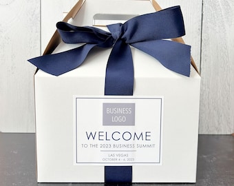 May include: A white gift box with a blue ribbon tied around it. The box has a label that says "Welcome to the 2023 Business Summit" and "Las Vegas, October 4-6, 2023". There is a gray rectangle with the text "Business Logo" above the welcome message.