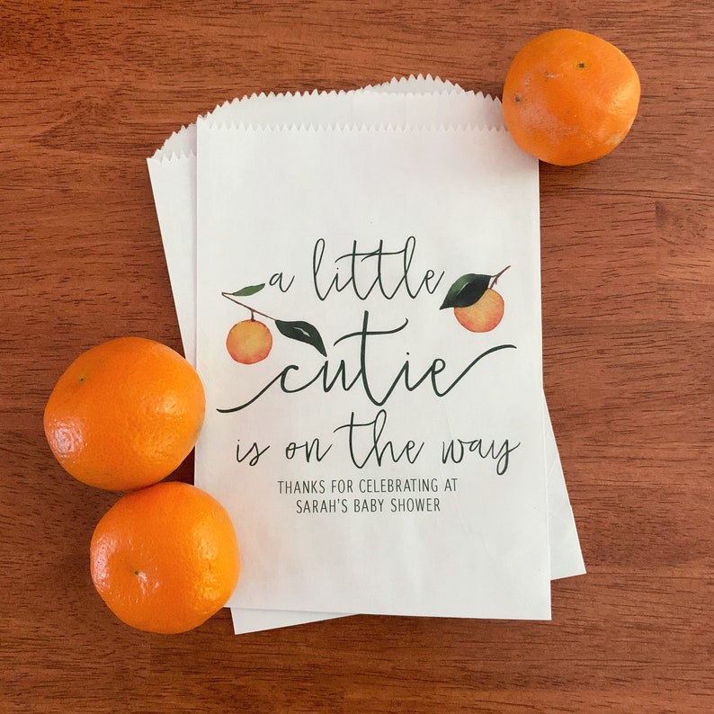 May include: White paper treat bags with a scalloped top, featuring the text "a little cutie is on the way" with orange illustrations. Three oranges are placed around the bags. The text also says "Thanks for celebrating at Sarah's baby shower."