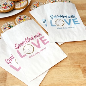 May include: Two white paper bags with pink and blue designs. The bags have a donut with sprinkles and the text "Sprinkled with LOVE" and "Katie's Baby Shower".