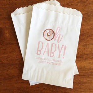 May include: White paper favor bags with pink text that reads "Oh Baby!" and a donut illustration. The text below the donut reads "Thanks for celebrating at Jenny's Baby Shower!"