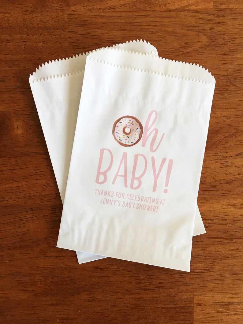 May include: White paper favor bags with pink text that reads "Oh Baby!" and a donut illustration. The text below the donut reads "Thanks for celebrating at Jenny's Baby Shower!"