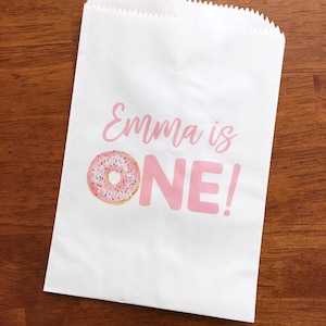 K&ouml;nnte beinhalten: Ein wei&szlig;er Papier-Geschenkbeutel mit rosa Schriftzug "Emma ist EIN!" und einer Illustration eines rosafarbenen und wei&szlig;en Donuts.
