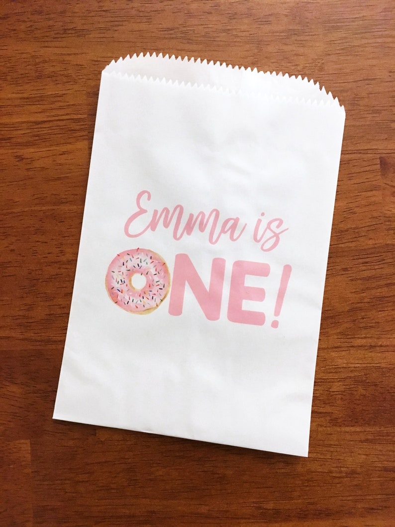 K&ouml;nnte beinhalten: Ein wei&szlig;er Papier-Geschenkbeutel mit rosa Schriftzug "Emma ist EIN!" und einer Illustration eines rosafarbenen und wei&szlig;en Donuts.