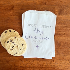 May include: White paper favor bag with the text "Thanks for celebrating my Holy Communion Olivia Smith April 2, 2022" and a purple cross.