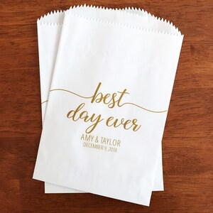 May include: Two white paper treat bags with a gold script that reads "best day ever." Below the text are the names "Amy & Taylor" and the date "December 9, 2018." The bags are on a dark wood surface.