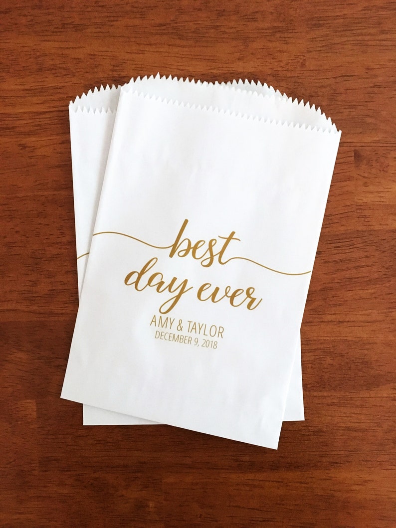 May include: Two white paper treat bags with a gold script that reads "best day ever." Below the text are the names "Amy & Taylor" and the date "December 9, 2018." The bags are on a dark wood surface.