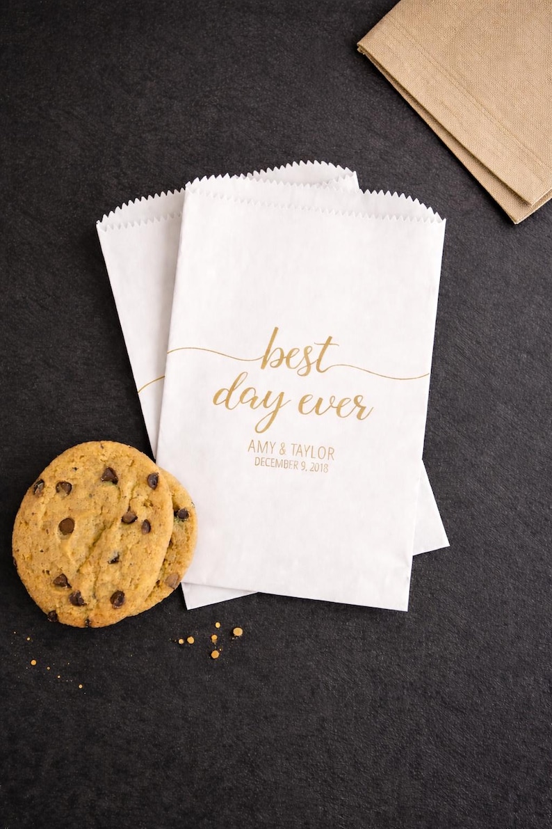 May include: White paper treat bags with a scalloped top, printed with "best day ever" in gold script, along with a date and names. Two chocolate chip cookies are next to the bags. A brown napkin is in the background.