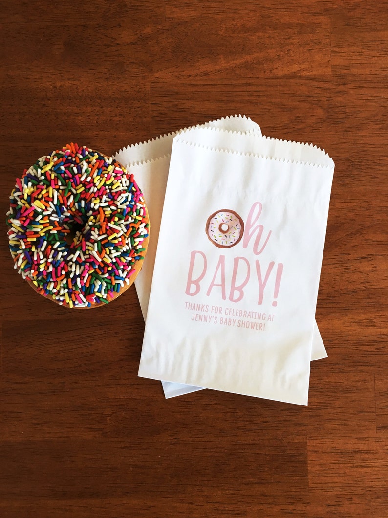 May include: White paper favor bags with pink text that reads "Oh Baby!" and a pink donut with sprinkles. The text below the donut reads "Thanks for celebrating at Jenny's Baby Shower!"