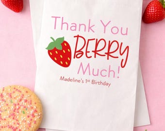 May include: A white paper favor bag featuring a red strawberry illustration and the text Thank You BERRY Much! Madeline's 1st Birthday in pink and red lettering. The bag is surrounded by fresh strawberries and a round cookie with sprinkles.