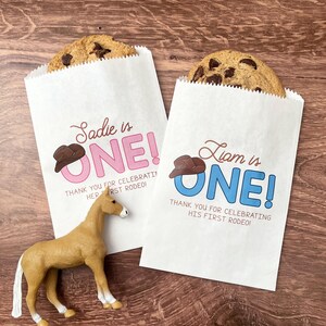 Peut inclure: Deux sacs en papier blanc avec un motif de chapeau de cowboy et le texte "Sadie is ONE!" et "Liam is ONE!"  Les sacs sont remplis de biscuits aux pépites de chocolat.  Le texte sur les sacs dit "Merci d'avoir célébré son/sa premier rodéo !"