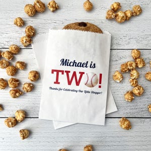May include: White paper treat bags with the text "Michael is TWO!" in navy and red, with a baseball graphic. A chocolate chip cookie peeks out of the top of one bag. Caramel popcorn surrounds the bags on a light wood surface.