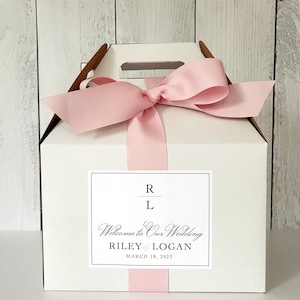 May include: White gift box with a pink ribbon and bow. The box has a label that says "Welcome to Our Wedding Riley & Logan March 18, 2023".