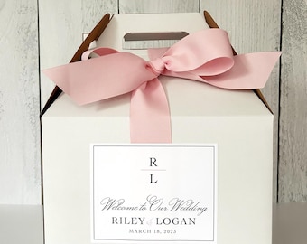 May include: White gift box with a pink ribbon and bow. The box has a label that says "Welcome to Our Wedding Riley & Logan March 18, 2023".