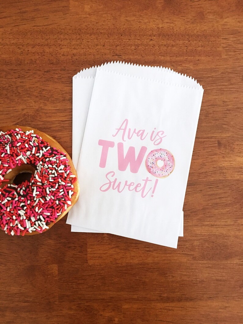 May include: Two white paper treat bags with pink text that reads "Ava is TWO Sweet!" and a donut graphic. A glazed donut with pink and red sprinkles sits to the left on a wooden surface.