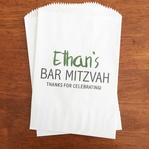 May include: Two white paper favor bags with a scalloped top edge. The bags have the text "Ethan's BAR MITZVAH THANKS FOR CELEBRATING!" printed in green and black. The bags are on a brown wooden surface.