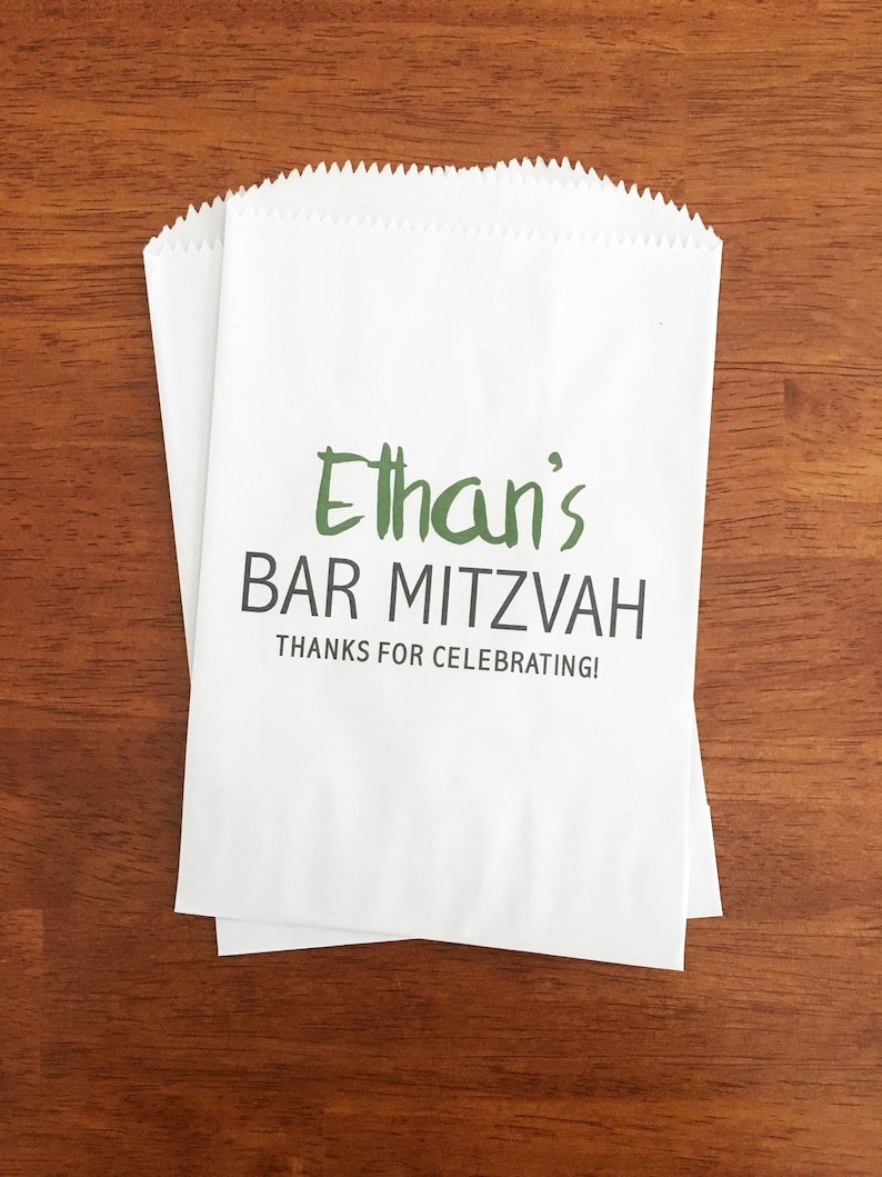 May include: Two white paper favor bags with a scalloped top edge. The bags have the text "Ethan's BAR MITZVAH THANKS FOR CELEBRATING!" printed in green and black. The bags are on a brown wooden surface.