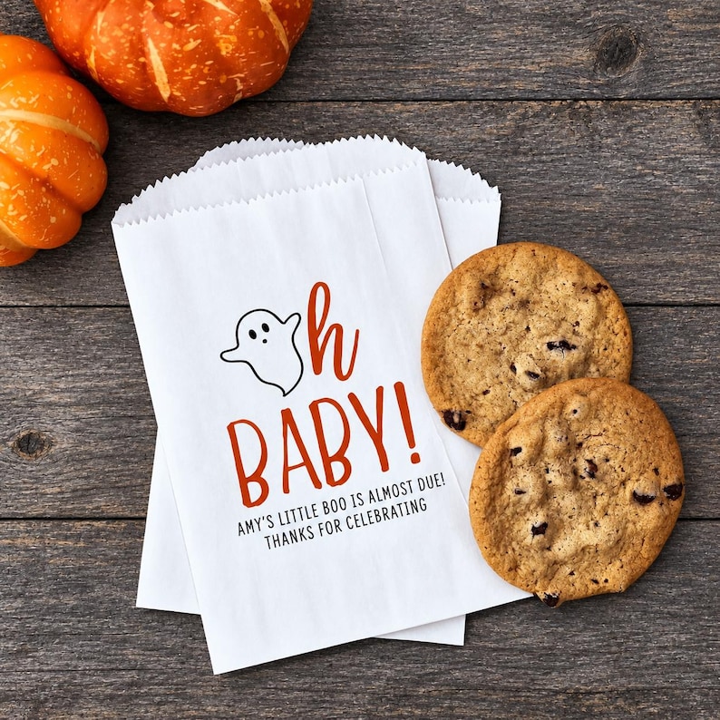 May include: A white paper treat bag featuring a black ghost illustration, the orange text "oh BABY!", and the black text "AMY'S LITTLE BOO IS ALMOST DUE! THANKS FOR CELEBRATING". Two round chocolate chip cookies are placed next to the paper bag.