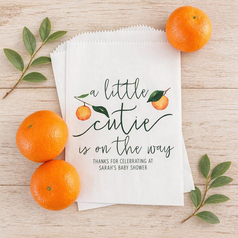 May include: White paper treat bags with the text "a little cutie is on the way" and orange illustrations. Three oranges and green sprigs are arranged around the bags on a light wood surface. The bags are for Sarah's baby shower.