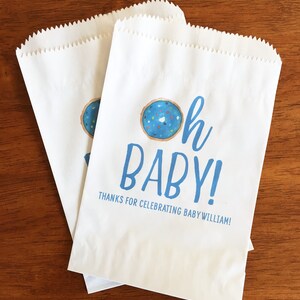 May include: Two white paper bags with blue and white text that reads "Oh Baby!" and "Thanks for celebrating Baby William!"  A blue and white donut with sprinkles is above the text.