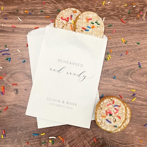 May include: White paper favor bags with the text "REHEARSED and ready" and "OLIVIA & MARK OCTOBER 3, 2024". The bags are filled with cookies topped with colorful sprinkles.