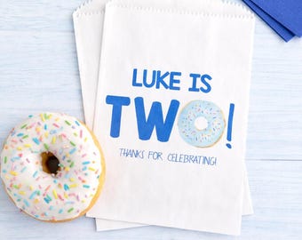 May include: White paper treat bags with blue text that reads "Luke is Two!" and a donut graphic. A frosted donut with colorful sprinkles sits next to the bags. Blue napkins are in the background.