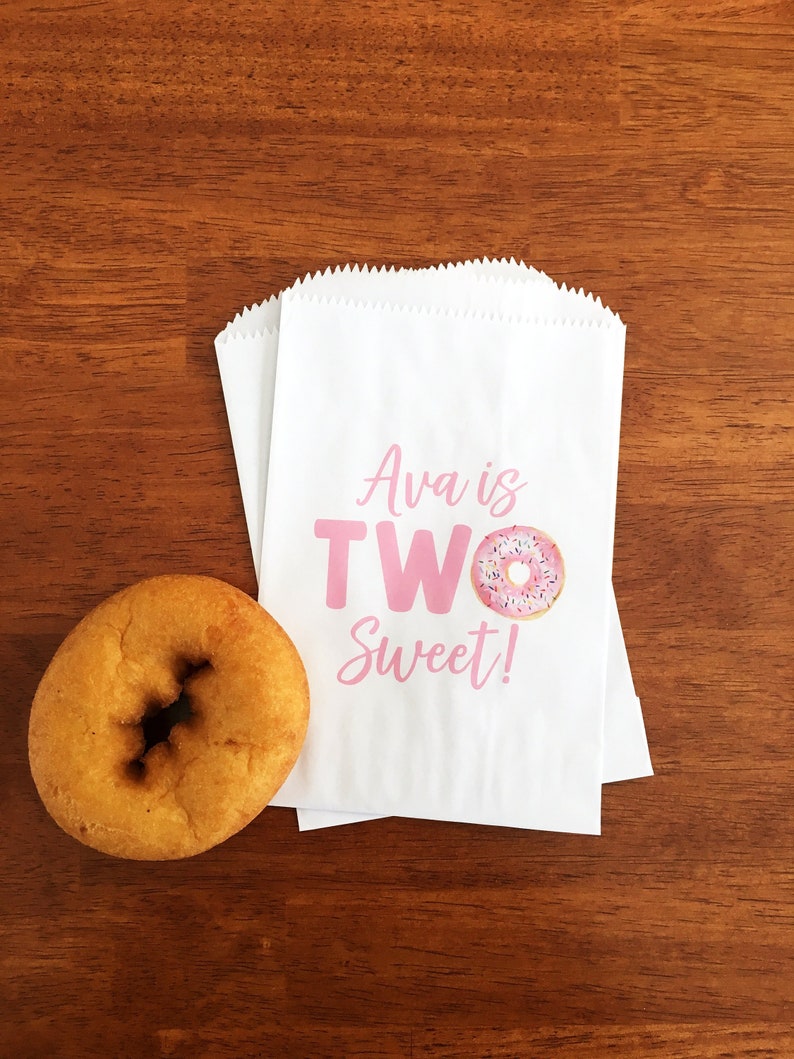 May include: A golden-brown donut sits next to three white paper treat bags. The bags have a pink text that reads "Ava is TWO Sweet!" with a donut graphic. The bags are on a wooden surface.