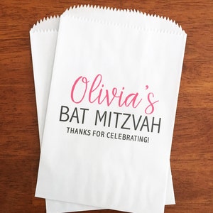 May include: Two white paper treat bags with a scalloped top edge. The bags have the text "Olivia's BAT MITZVAH Thanks for Celebrating!" in pink and black lettering.