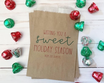 May include: A brown paper bag with the text "WISHING YOU A sweet HOLIDAY SEASON FROM THE DURYEA'S" printed in red and green. The bag is surrounded by scattered red, green, and silver foil-wrapped chocolate candies on a white wooden surface.