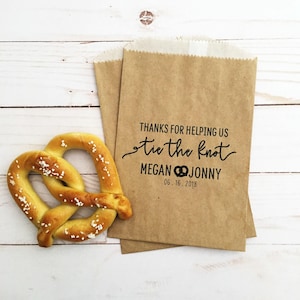 Peut inclure: Un sac en papier kraft brun imprimé avec le texte noir THANKS FOR HELPING US tie the knot MEGAN JONNY 06.16.2018, accompagné d'un bretzel salé doré. Le sac présente une petite icône de bretzel entre les prénoms.