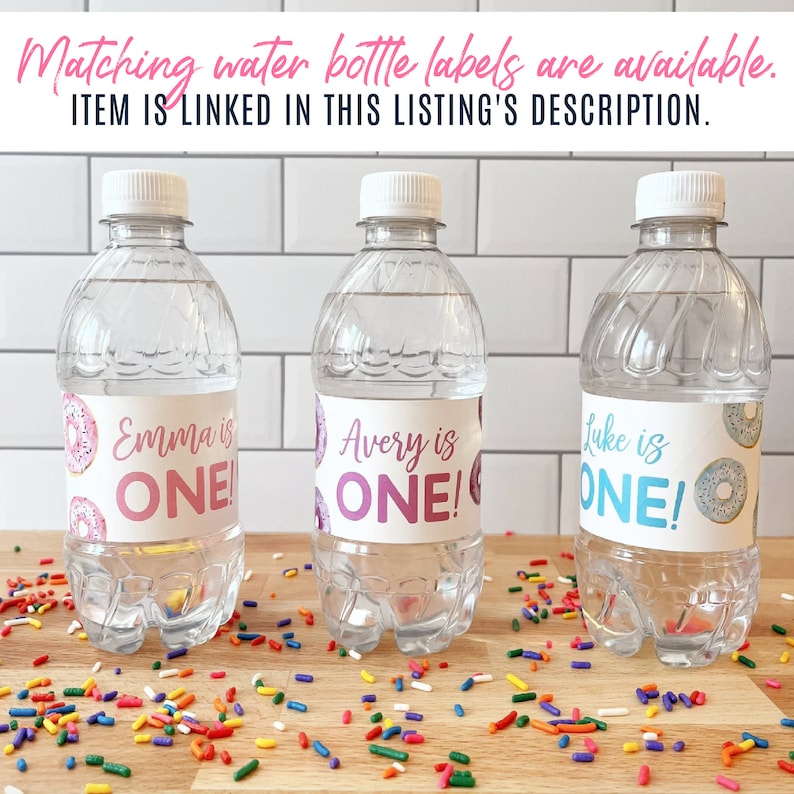 K&ouml;nnte beinhalten: Drei durchsichtige Plastikwasserflaschen mit personalisierten Etiketten f&uuml;r eine erste Geburtstagsfeier. Die Etiketten zeigen ein Donut-Design und den Text "Emma ist EINS!", "Avery ist EINS!" und "Luke ist EINS!"