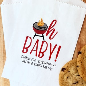 Może przedstawiać: Biała papierowa torba z grafiką grilla w kolorze czerwonym i czarnym oraz tekstem "Oh Baby!" w kolorze czerwonym. Pod grafiką znajduje się tekst "Dzięki za świętowanie u Olivii i Ryana na Baby-Q!"