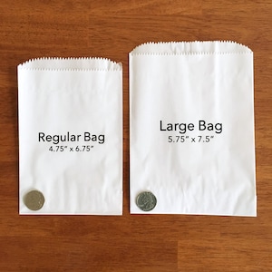 K&ouml;nnte beinhalten: Zwei wei&szlig;e Papiert&uuml;ten, eine mit der Aufschrift "Regular Bag 4.75" x 6.75"" und die andere mit der Aufschrift "Large Bag 5.75" x 7.5"". Eine US-amerikanische 25-Cent-M&uuml;nze liegt vor jeder T&uuml;te zur Gr&ouml;&szlig;envergleich.