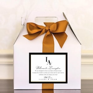 May include: A white gift box tied with a gold satin ribbon and bow. A rectangular label with black border is affixed to the front, displaying the text "Welcome to Lexington" and the names "Laura & Adam".