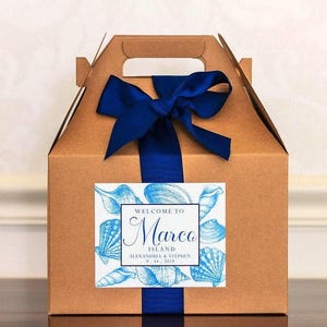 May include: A brown cardboard gift box with a blue ribbon and bow. The box has a label that says "Welcome to Marco Island, Alexandria & Stephen 9.14.2019". The label also features a blue and white illustration of seashells.