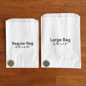 May include: Two white paper bags, labeled "Regular Bag" and "Large Bag." The regular bag measures 4.75" x 6.75", and the large bag is 5.75" x 7.5". A coin is placed next to each bag for size comparison. The bags are on a wooden surface.