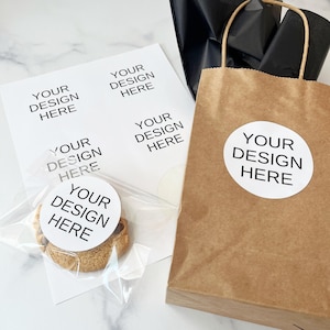 May include: A brown paper bag with a white circle sticker that says "YOUR DESIGN HERE". The bag is filled with black tissue paper. There are four white cards with the same text on them. One card is in a clear plastic bag with a chocolate chip cookie with a white circle sticker that says "YOUR DESIGN HERE".
