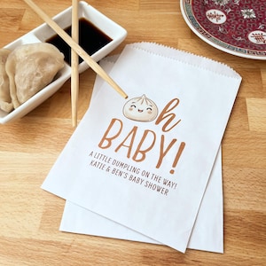 May include: White paper bag with a brown graphic of a dumpling with a smiling face and the text "Oh Baby! A little dumpling on the way! Katie & Ben's Baby Shower"