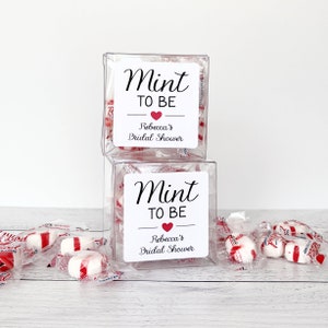 May include: Two clear plastic boxes filled with red and white peppermint candies. The boxes have white labels with black text that reads "Mint to Be" and "Rebecca's Bridal Shower".