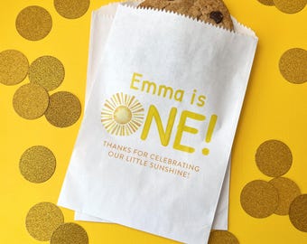 May include: White paper party favor bag with a yellow sun and the text "Emma is ONE!" and "Thanks for celebrating our little sunshine!"