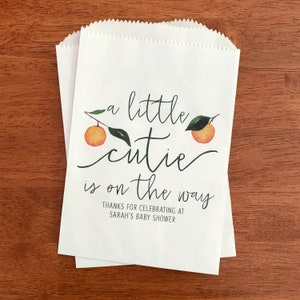 May include: Two white paper treat bags with a scalloped top. The bags feature the text "a little cutie is on the way" with orange and green watercolor illustrations. The text "Thanks for celebrating at Sarah's baby shower" is also included.
