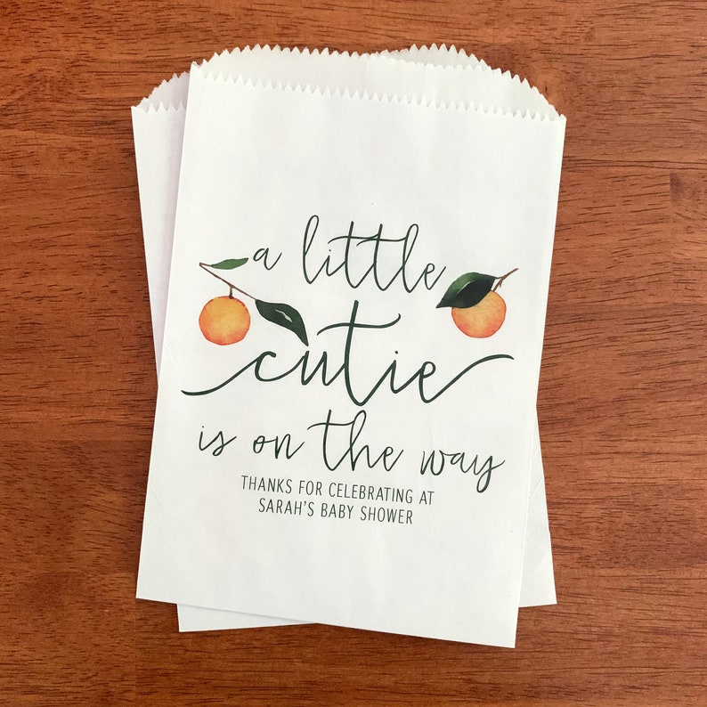May include: Two white paper treat bags with a scalloped top. The bags feature the text "a little cutie is on the way" with orange and green watercolor illustrations. The text "Thanks for celebrating at Sarah's baby shower" is also included.