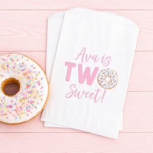May include: White paper treat bags with pink text that reads "Ava is TWO Sweet!" and a donut graphic. A frosted donut with colorful sprinkles sits next to the bags on a pink wooden surface.