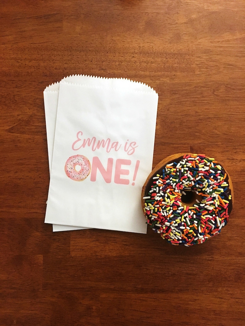 K&ouml;nnte beinhalten: Eine wei&szlig;e Papiert&uuml;te mit dem Text "Emma is ONE!" und einer Illustration eines rosafarbenen Donuts. Neben der T&uuml;te liegt ein Schokoladendonut mit bunten Streuseln.