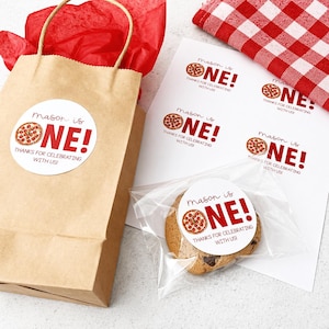 May include: A brown paper gift bag with a red tissue paper lining and a round sticker that says "Mason is ONE!" with a pizza graphic. A cookie in a clear bag has a matching sticker. Additional stickers are on a white sheet.