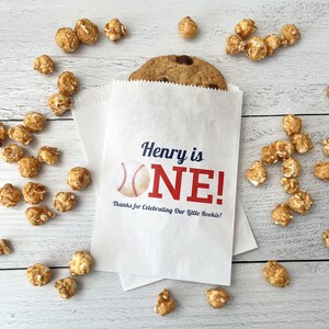 May include: A white paper bag with the text "Henry is ONE!" and a baseball graphic. A large chocolate chip cookie peeks out of the bag, surrounded by caramel popcorn. The text "Thanks for Celebrating Our Little Rookie!" is also printed on the bag.