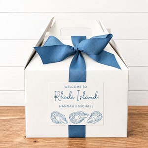 May include: A white gift box tied with a blue ribbon and bow. A rectangular label on the front reads "Welcome to Rhode Island" with the names "Hannah & Michael" below, and three oyster illustrations.