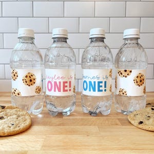 May include: Four clear plastic water bottles with personalised labels. The labels feature a cookie design and the text "[Name] is ONE!" in pink and blue.